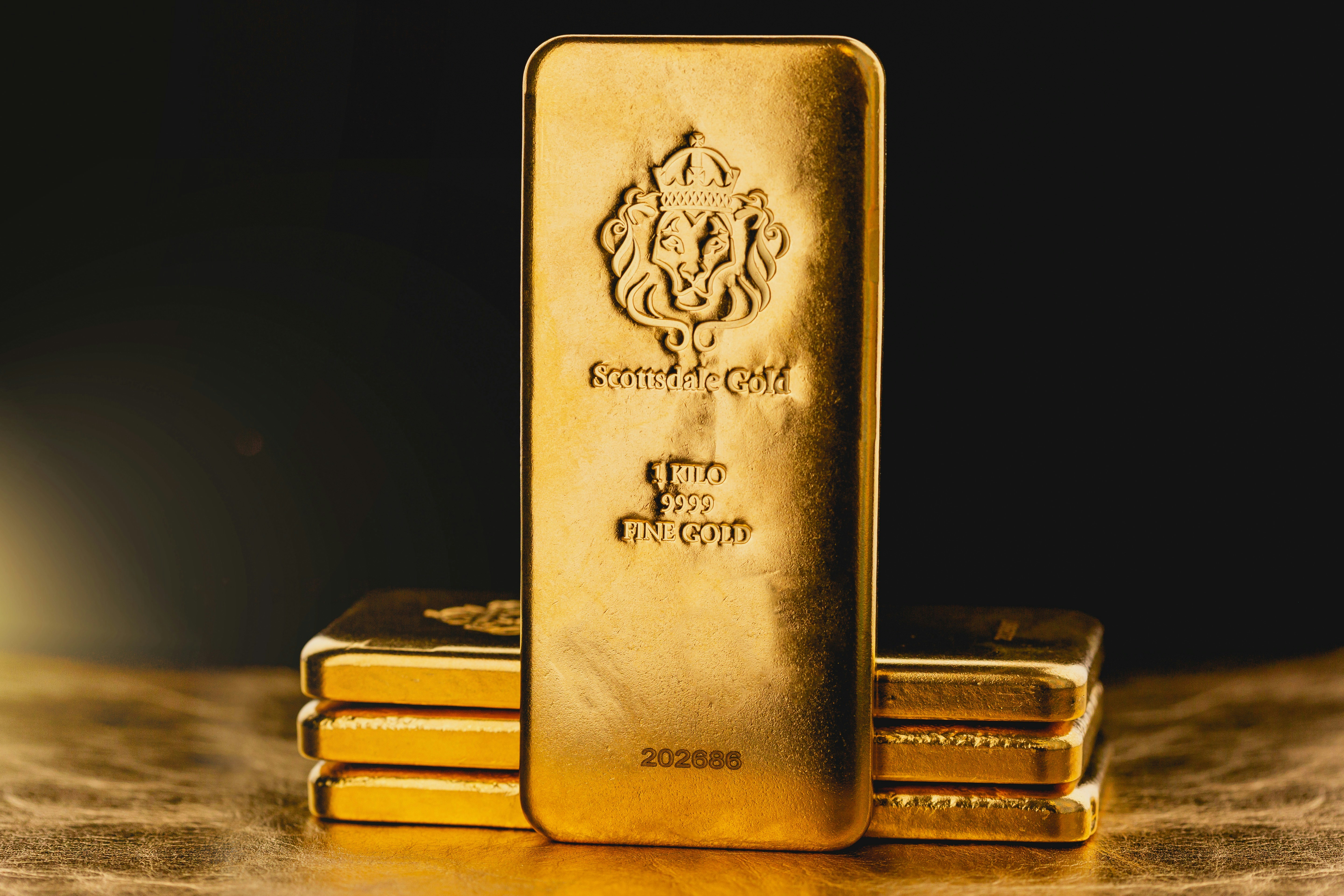 A stack of gold bars is prominently displayed. The forefront features a larger gold bar with intricate engravings, including a lion emblem and inscriptions indicating its weight as one kilo of fine gold. The scene is set against a dark background with a warm, golden glow, highlighting the richness and value of the bars.