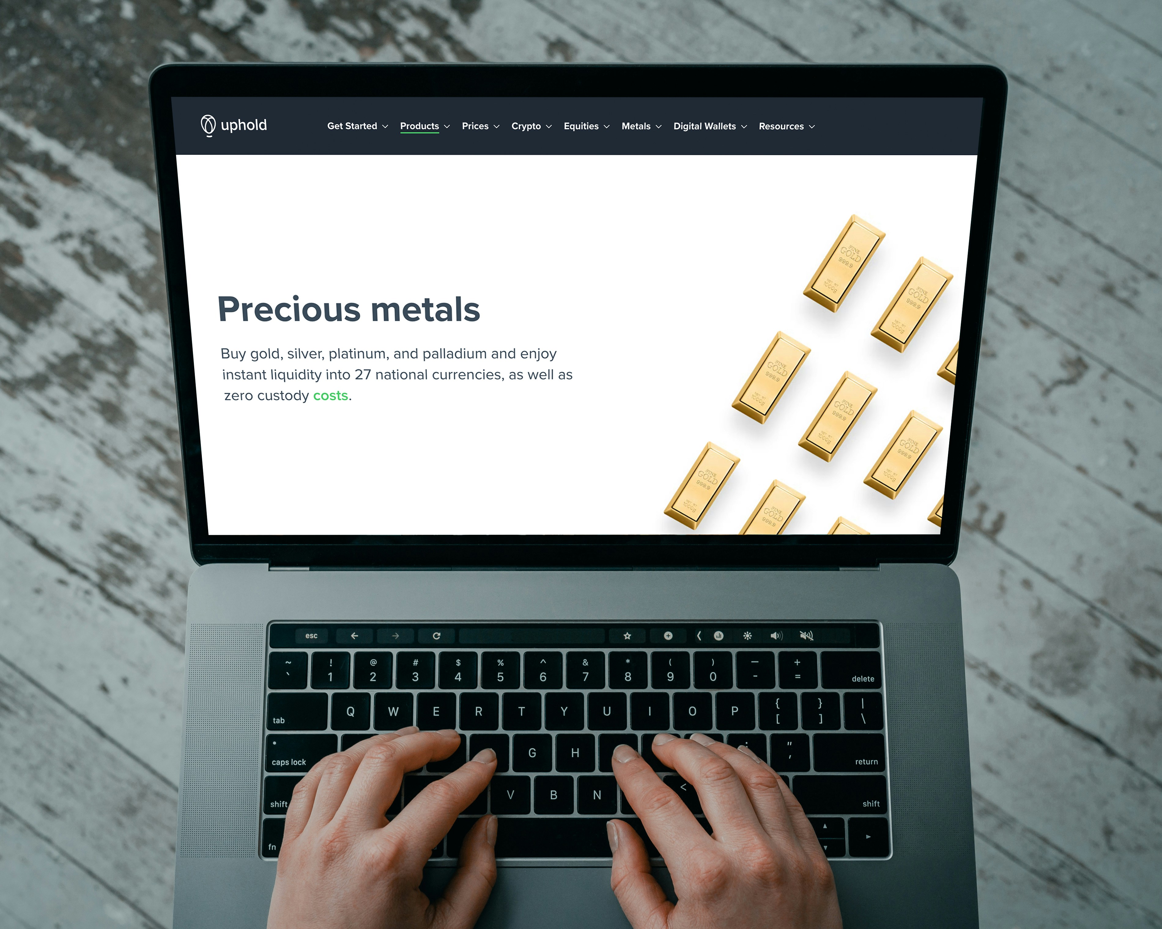 Hands typing on a laptop keyboard with an open webpage displaying information about buying precious metals such as gold, silver, platinum, and palladium. The screen shows an image of gold bars arranged in a pattern. The background consists of a wooden floor with a distressed look.